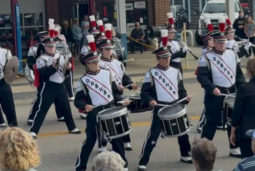 Marching band in black and white uniforms, playing drums and instruments in an outdoor parade setting.