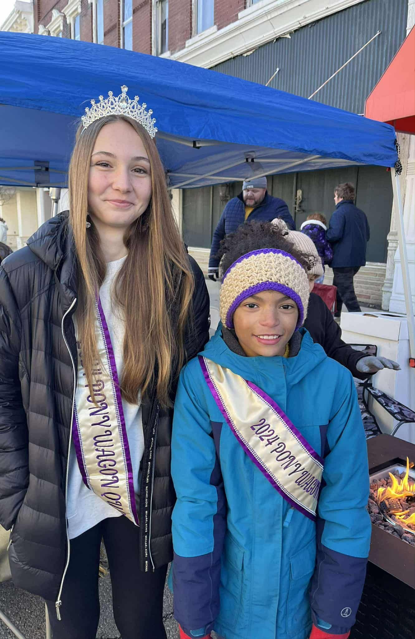 Two young people wearing sashes and a tiara, standing outdoors near a fire pit and a blue tent.