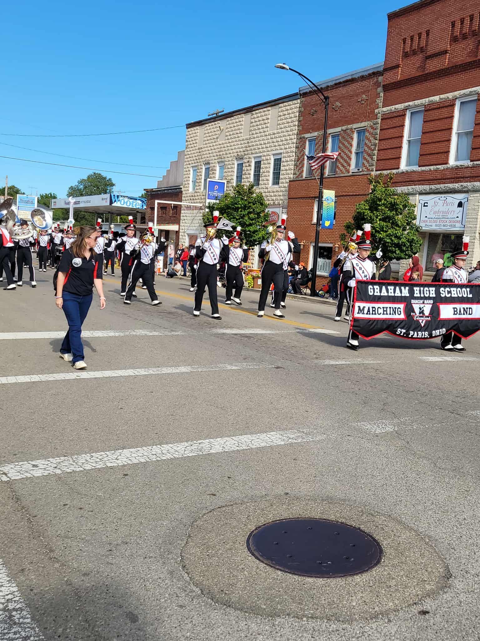 Marching band in black and white uniforms marching down a street in front of brick buildings on a sunny day.