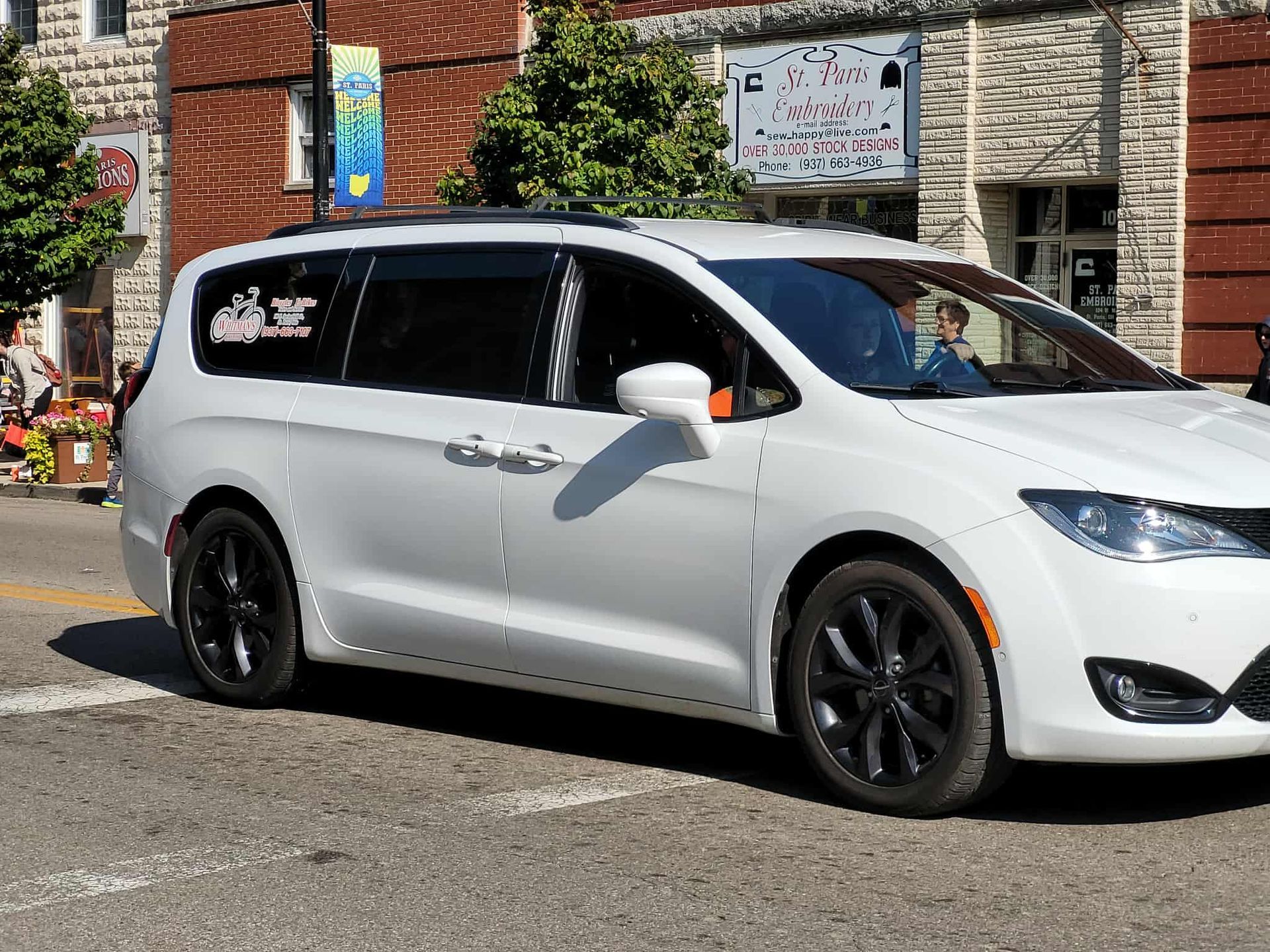 White minivan with black wheels driving on a street with a building in the background.