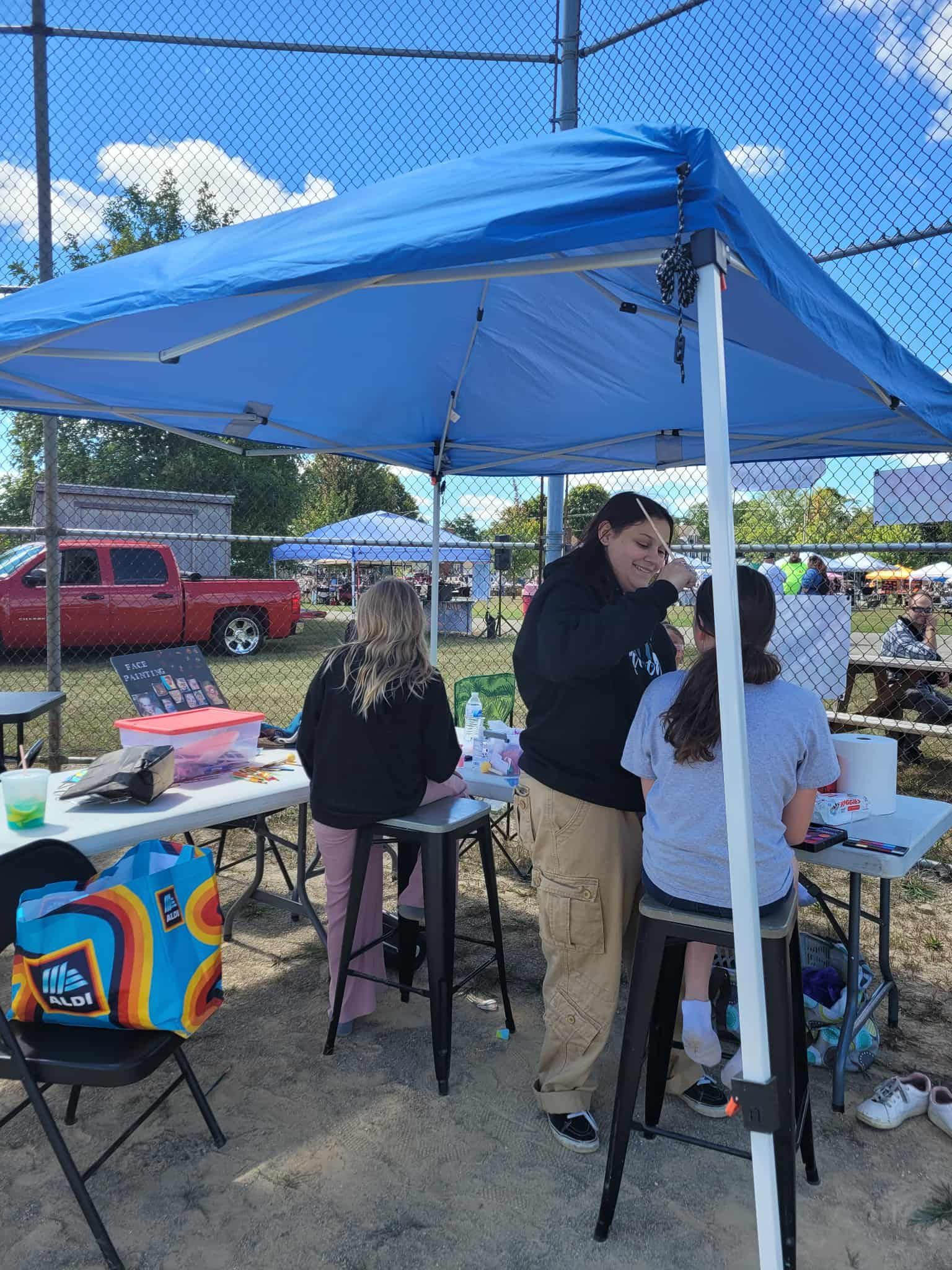 Face painting booth under a blue canopy on a sunny day. People are getting their faces painted.
