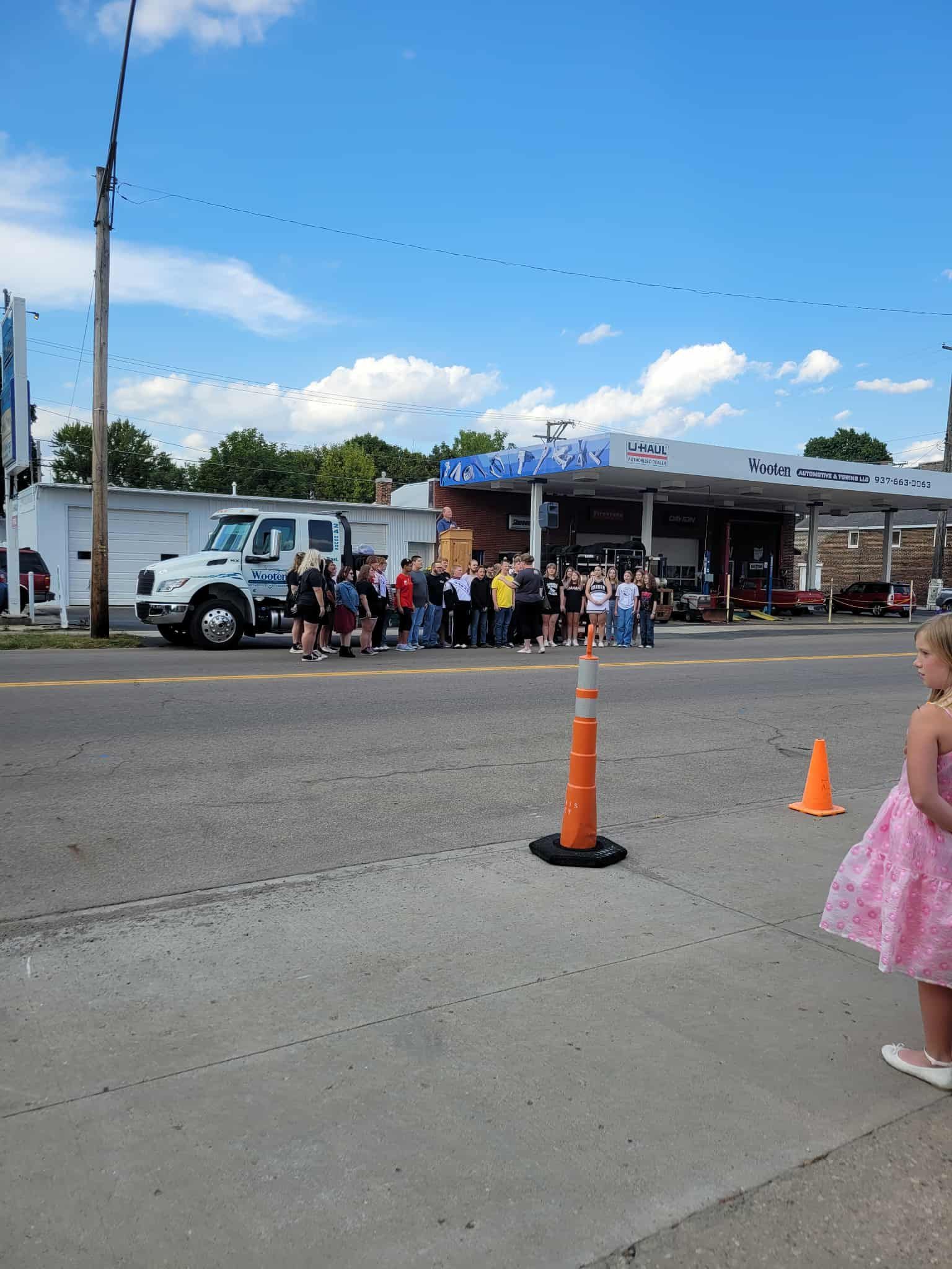 A crowd of people stands in front of a gas station under a blue sky, with a white truck and a girl in a pink dress in the foreground.