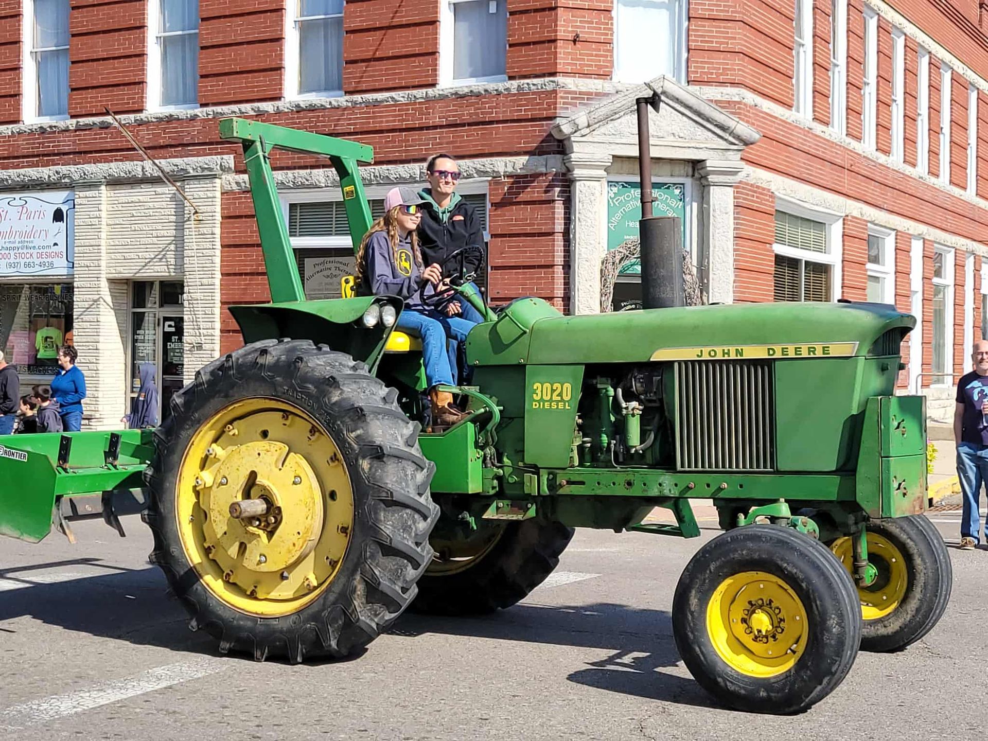 John Deere tractor with two people driving down a street with brick buildings.
