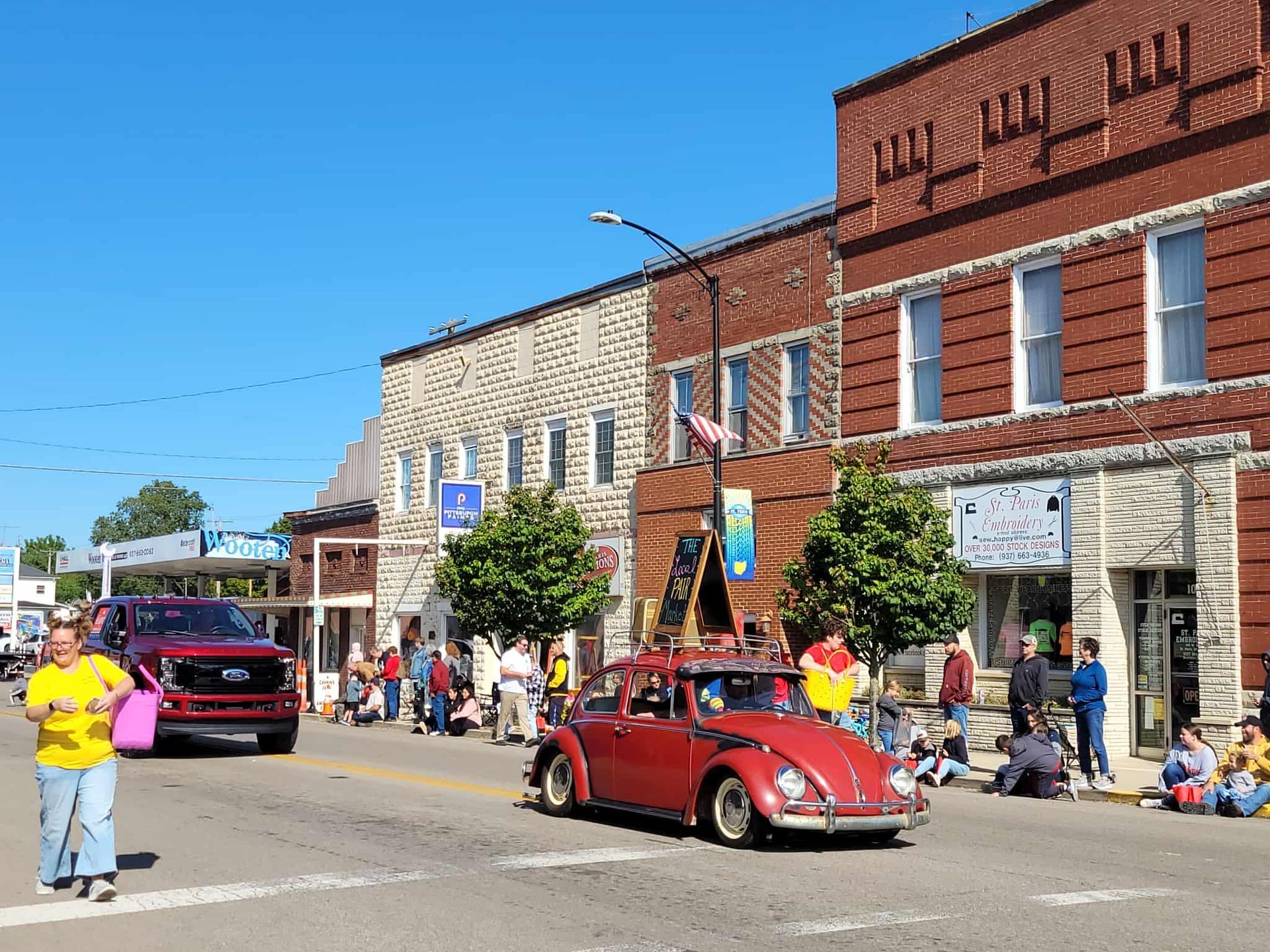 A classic red Volkswagen Beetle drives down a street during a parade. Buildings line the street with people watching.