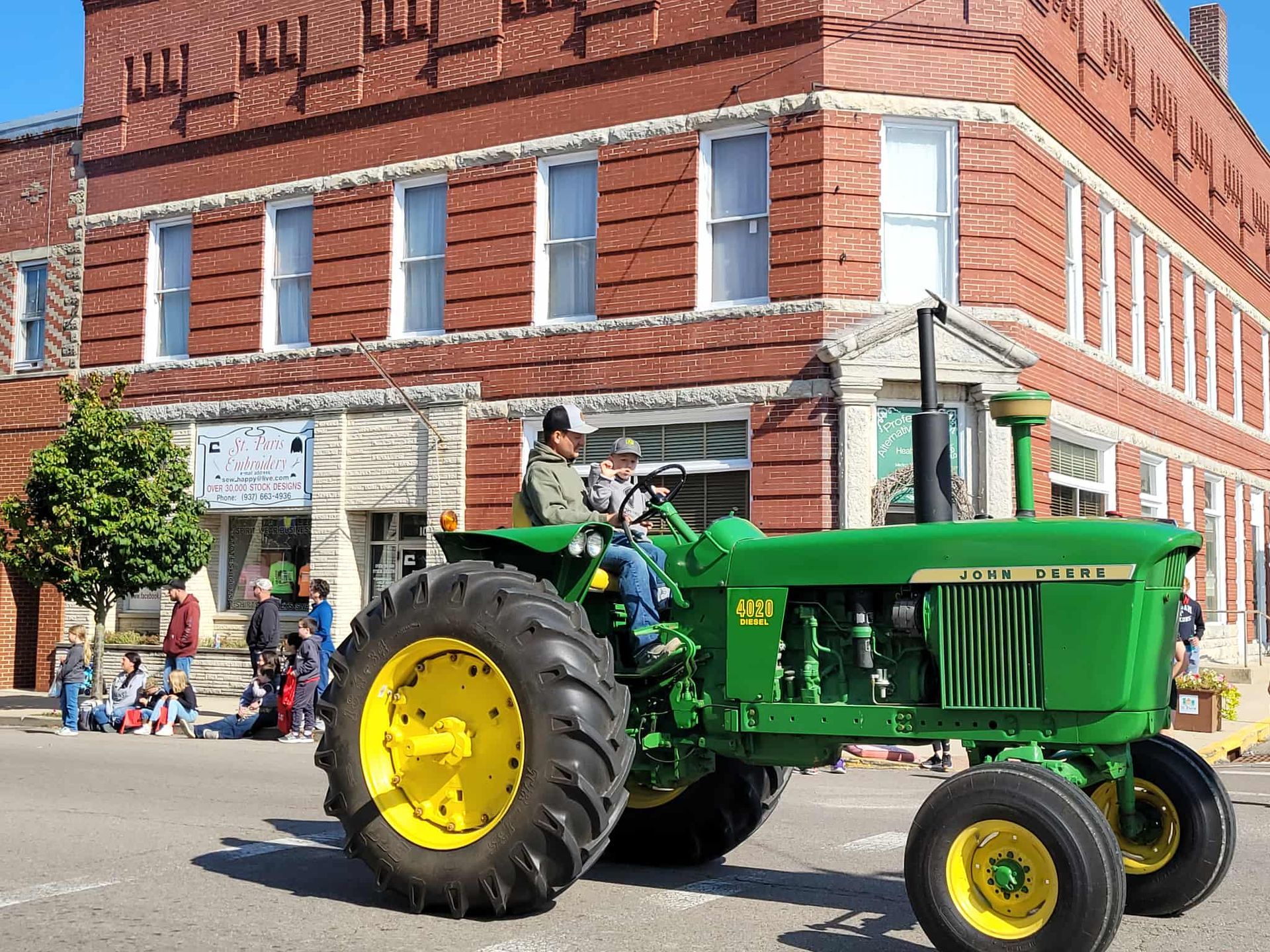 Green John Deere tractor with two people driving in a parade, brick building background.