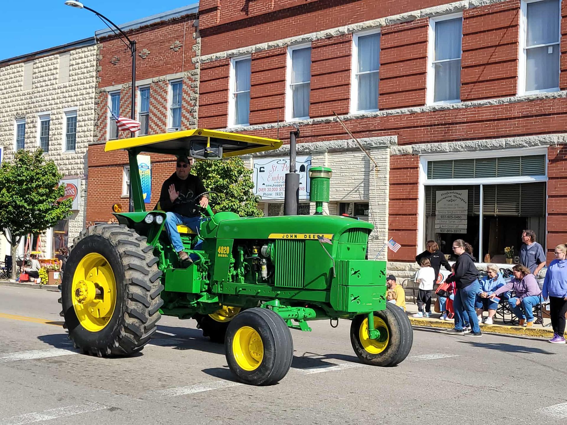 Green John Deere tractor in a parade, driver waving, people watching from sidewalk.