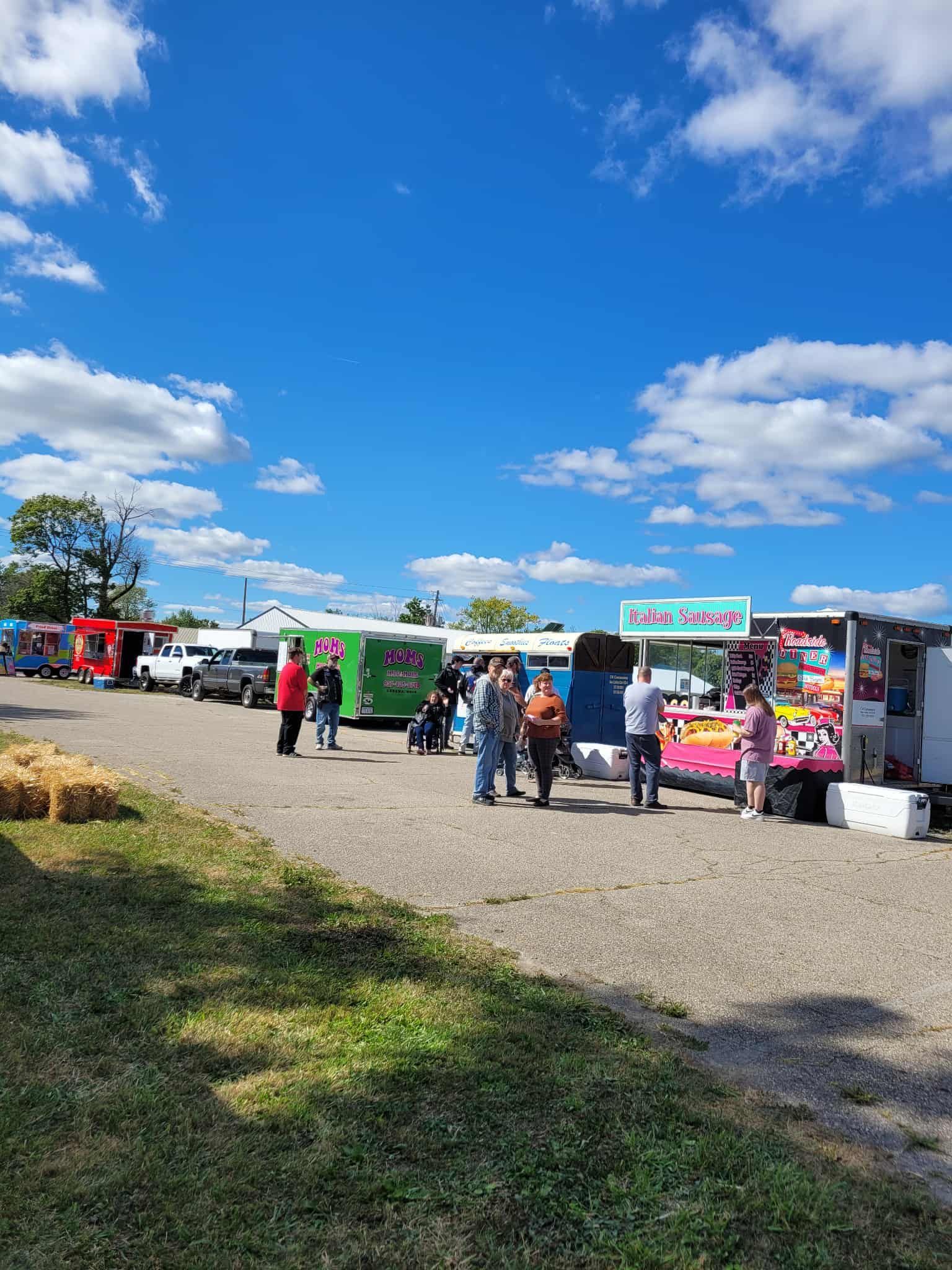 Food trucks and people at an outdoor event on a sunny day with blue sky and white clouds.