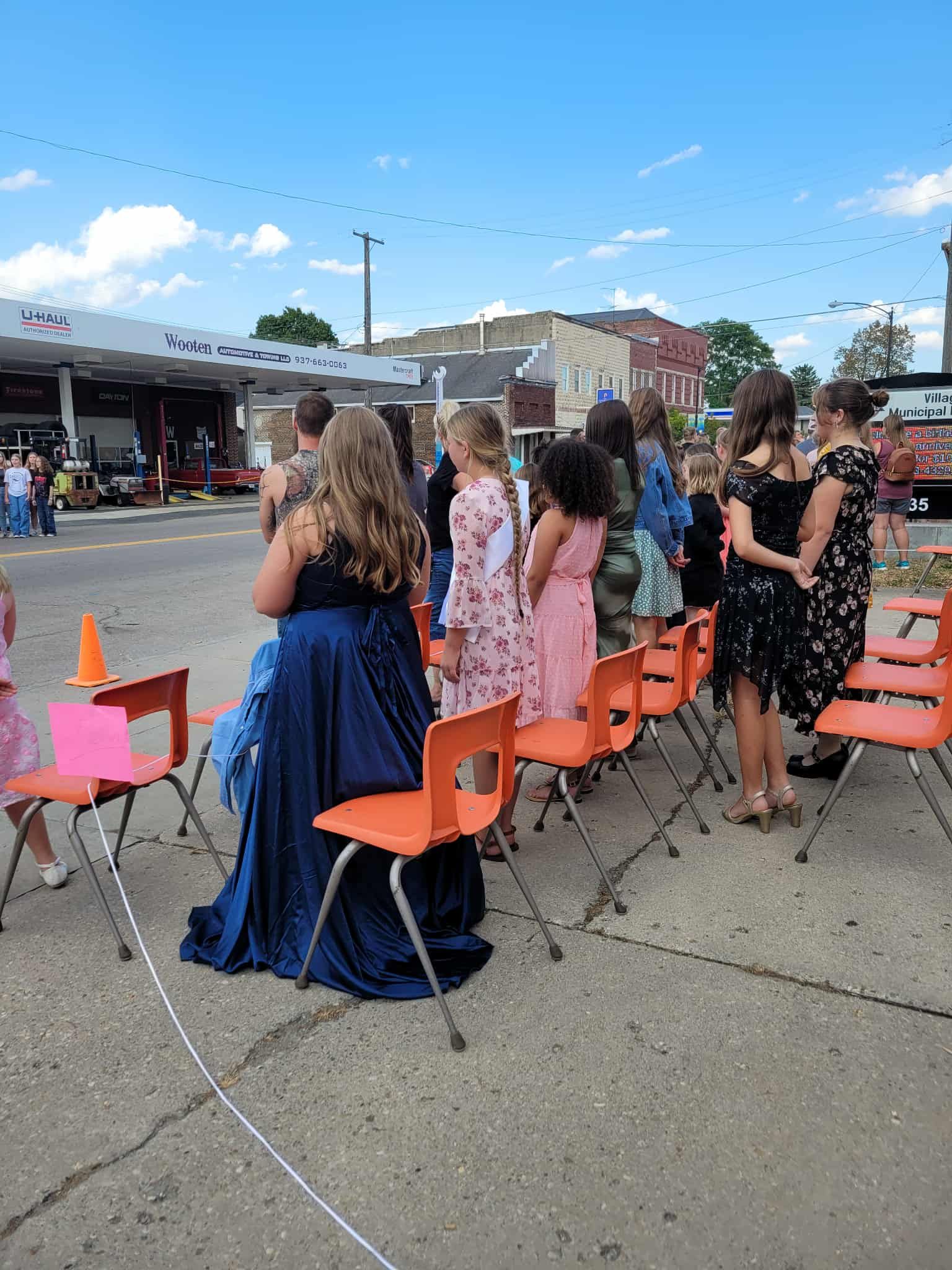 People in formal dresses stand in a row of orange chairs outside a gas station on a sunny day.