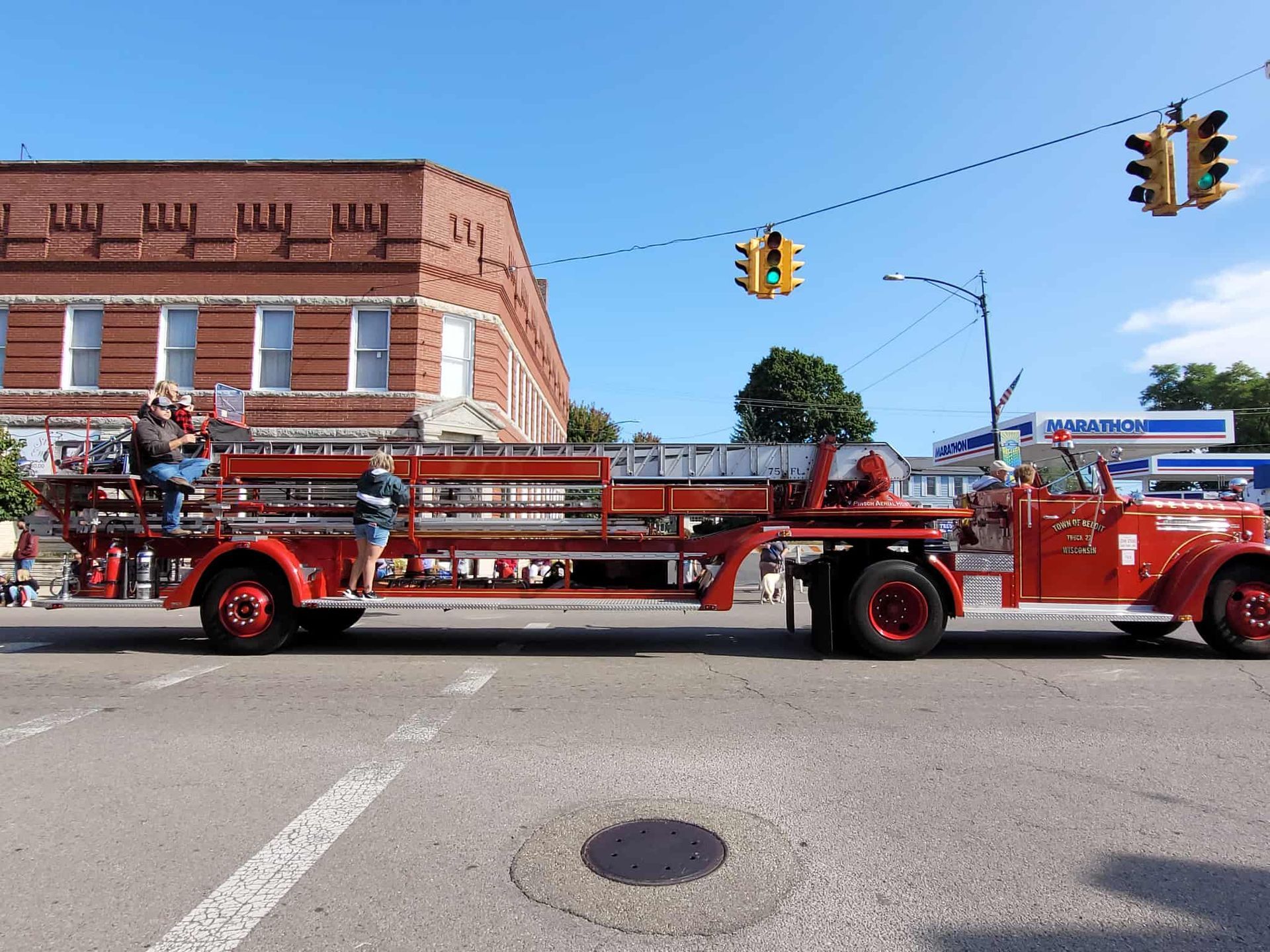 Red vintage fire truck with two people on a street, in front of a brick building and a traffic light.