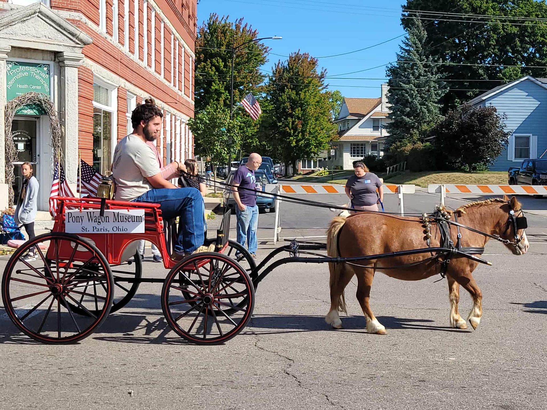 Man driving a horse-drawn cart down a street with spectators and buildings in the background.
