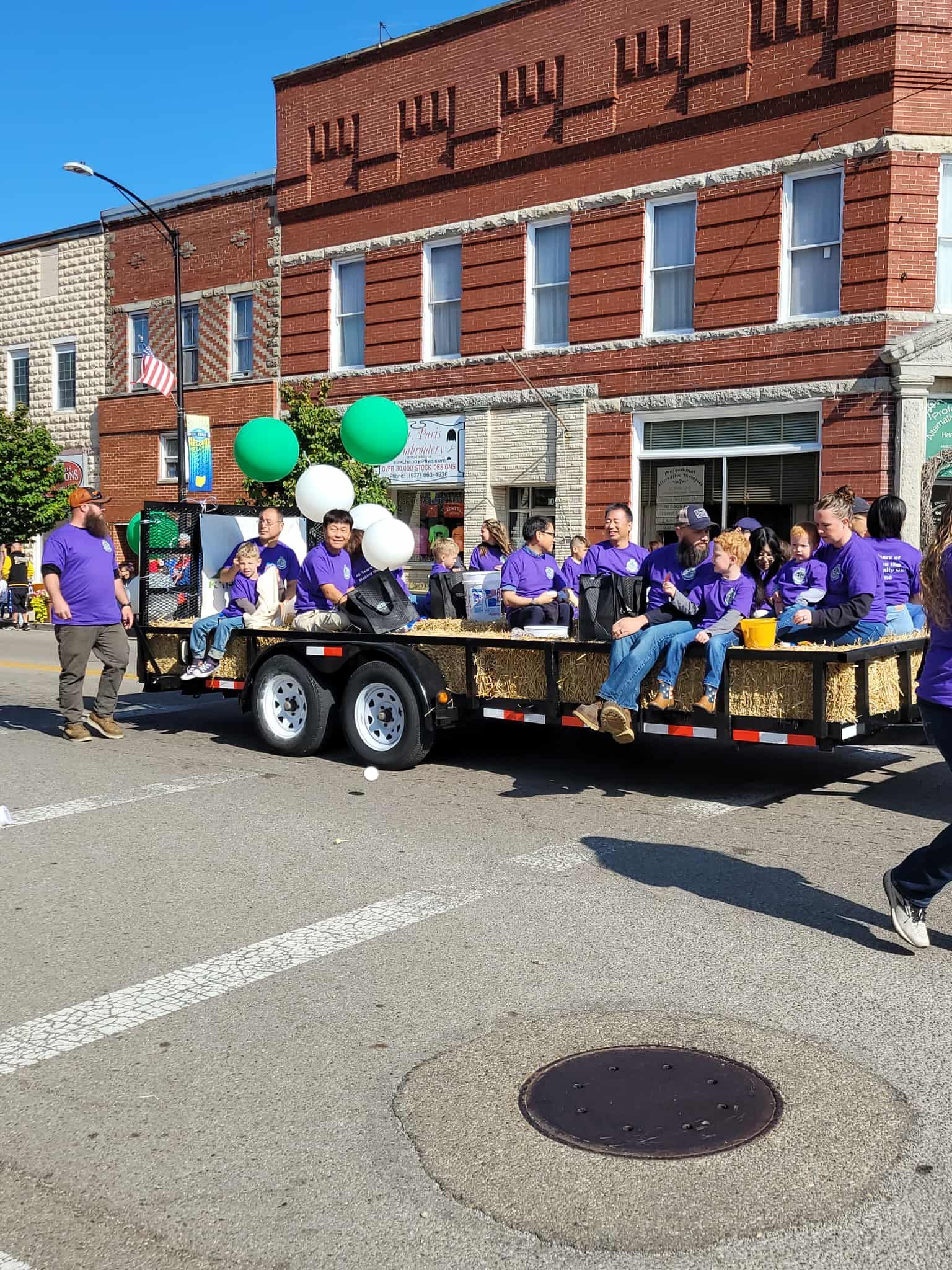 People on a float with green and white balloons, in front of a brick building.