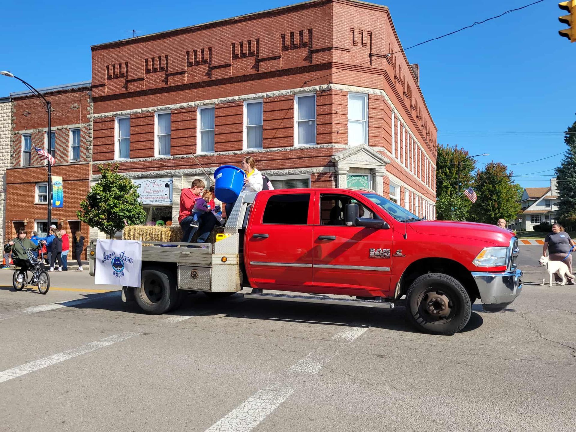 Red pickup truck with flatbed, people riding on hay bales, in a parade, brick building in the background.