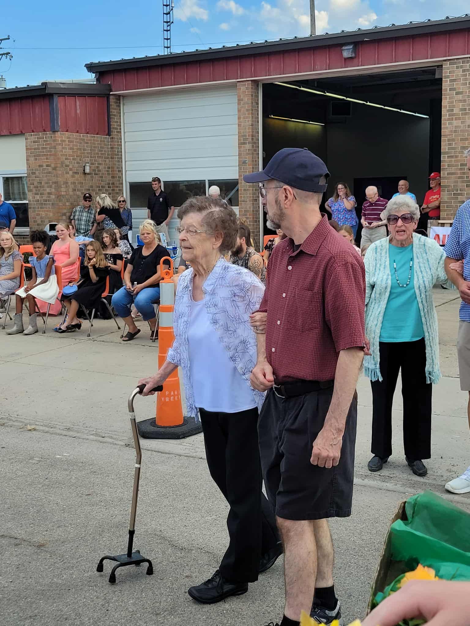 An older woman using a cane is helped by a man, walking near a building with a crowd.