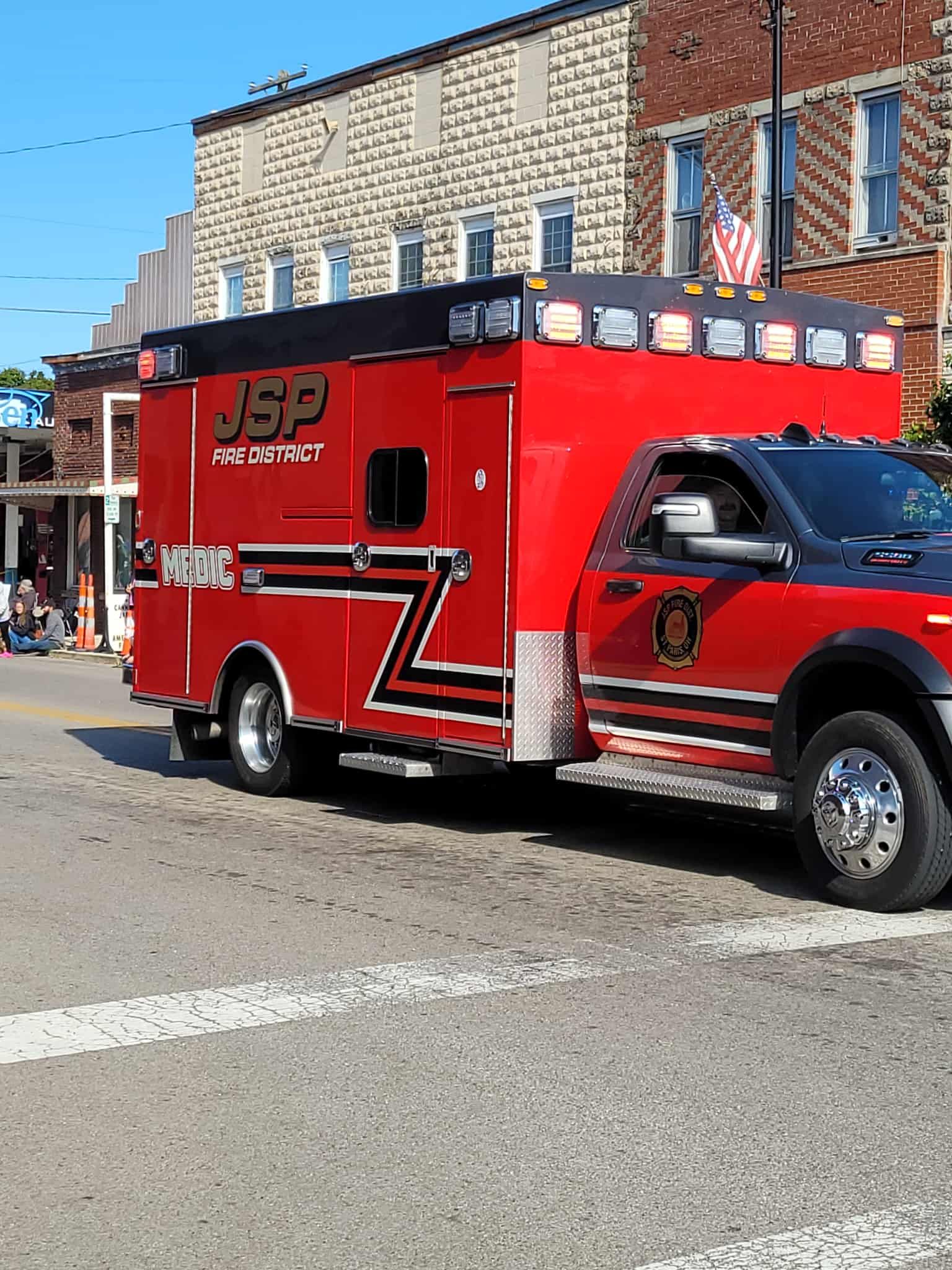 Red and black ambulance with flashing lights in a parade. JSP Grove on the side.