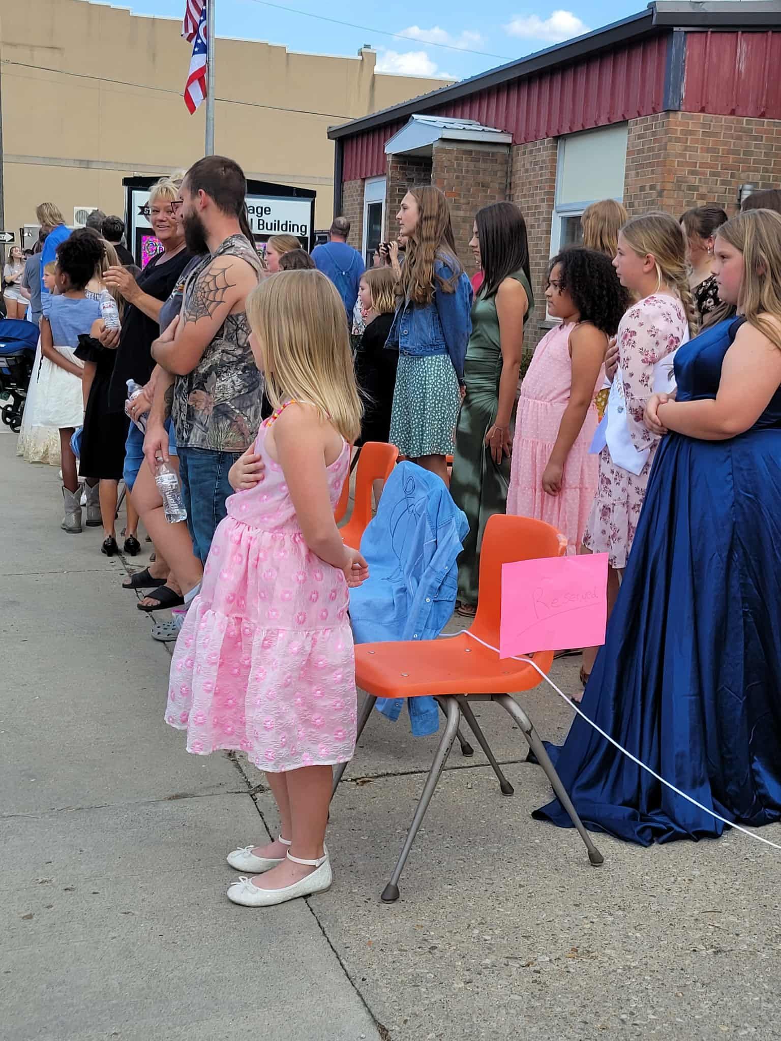 People line a street, some in formal dresses, near an orange chair with a pink sign, and buildings in the background.