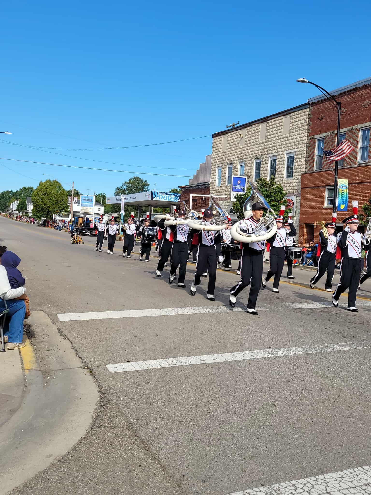 Marching band in black and white uniforms marching down a street in a town parade.