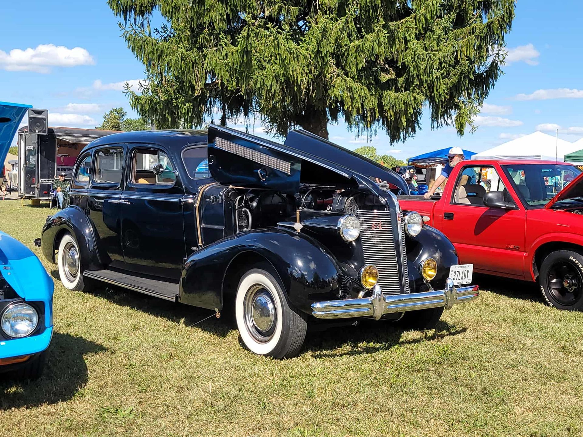 Black classic car with hood open, parked on grass at an outdoor event. White wall tires, red truck visible.