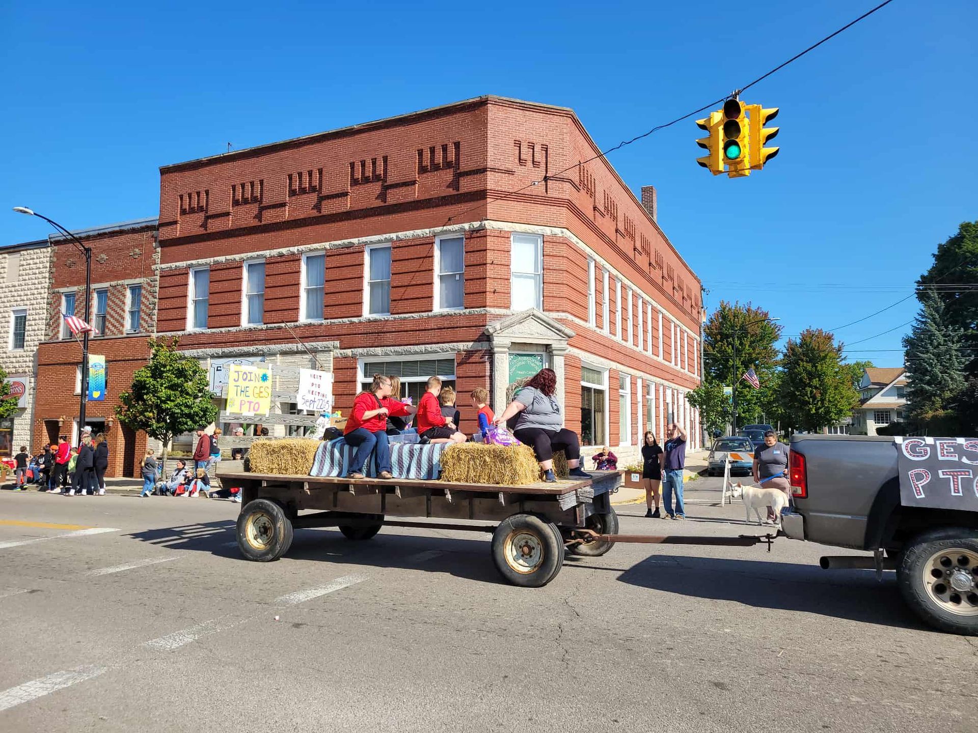 Parade float with people, hay bales, and red brick building on sunny street.