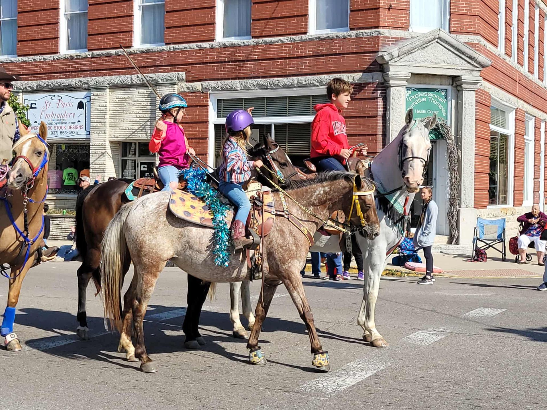 Children on horseback in a parade, riding past brick buildings.