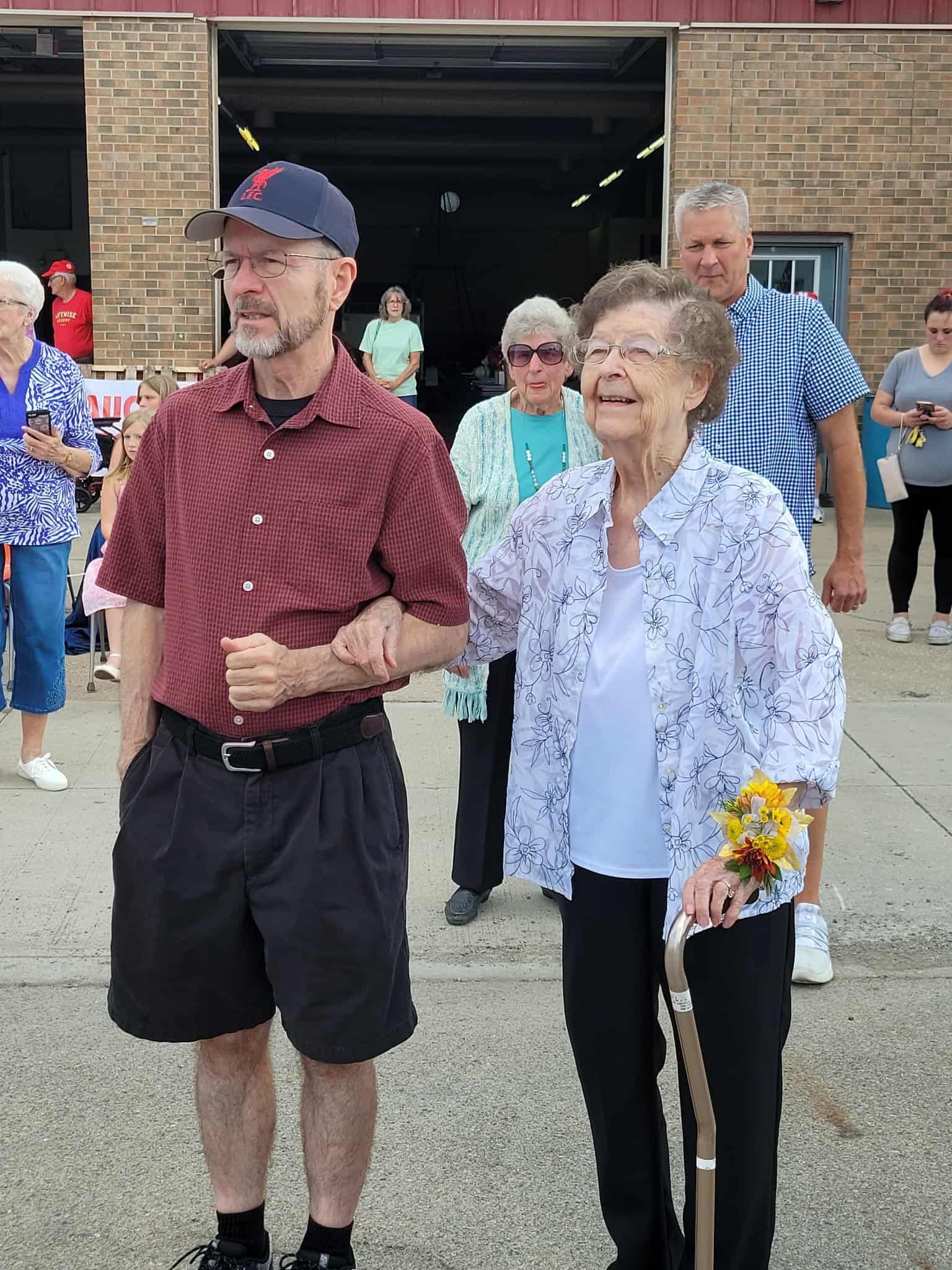 A man and a woman arm-in-arm outside a building; woman smiles holding a cane, people in background.
