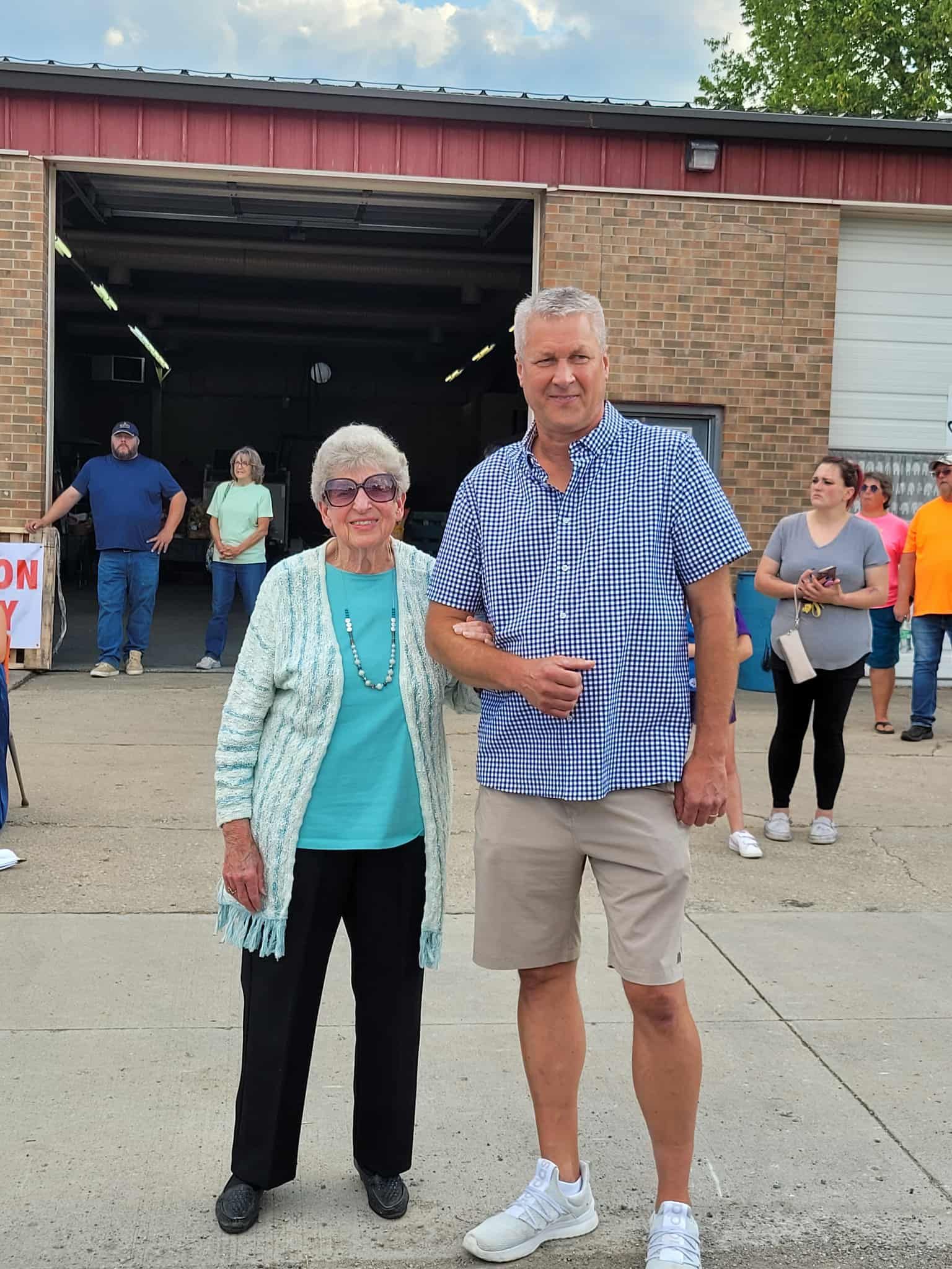 Man and woman standing in front of a building; man wearing a blue shirt and khaki shorts; woman in a cardigan.