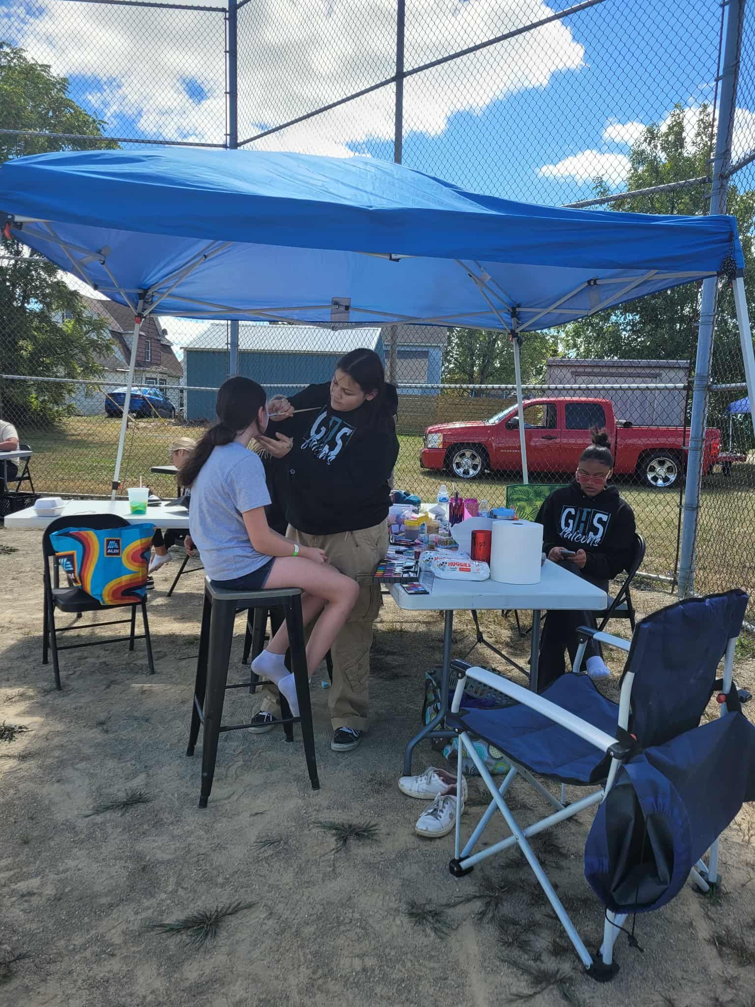 Face painting under a blue canopy at an outdoor event. A person paints a young girl's face; others sit nearby.