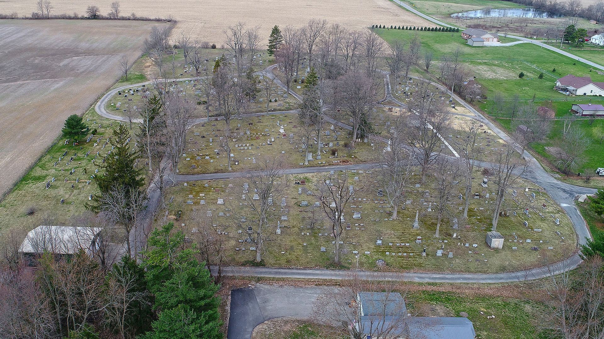 Aerial view of a cemetery with numerous headstones among bare trees. Paved paths wind through the grounds.