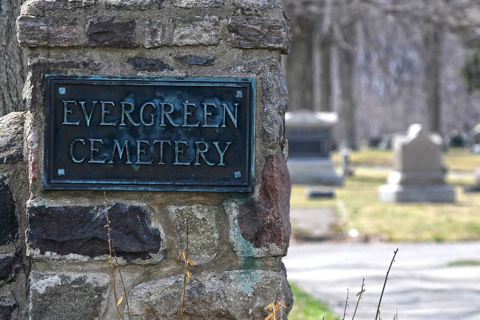 Sign for Evergreen Cemetery on a stone pillar, with tombstones in the background.