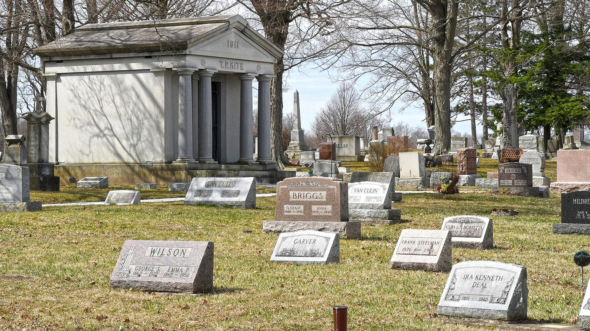 A cemetery with gravestones and a large mausoleum. Sunny day, dry grass, and trees.
