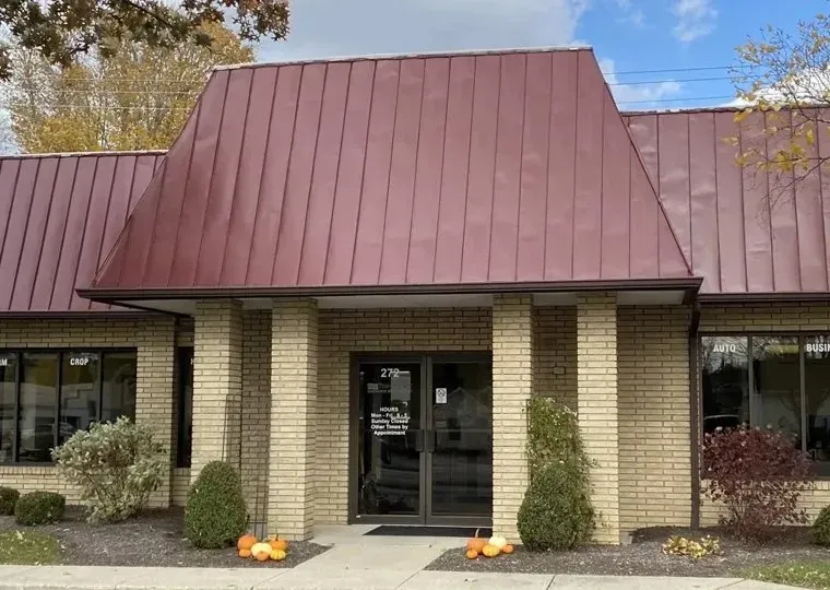 Brown brick building with a dark red metal roof, glass doors, and small bushes with pumpkins in front.