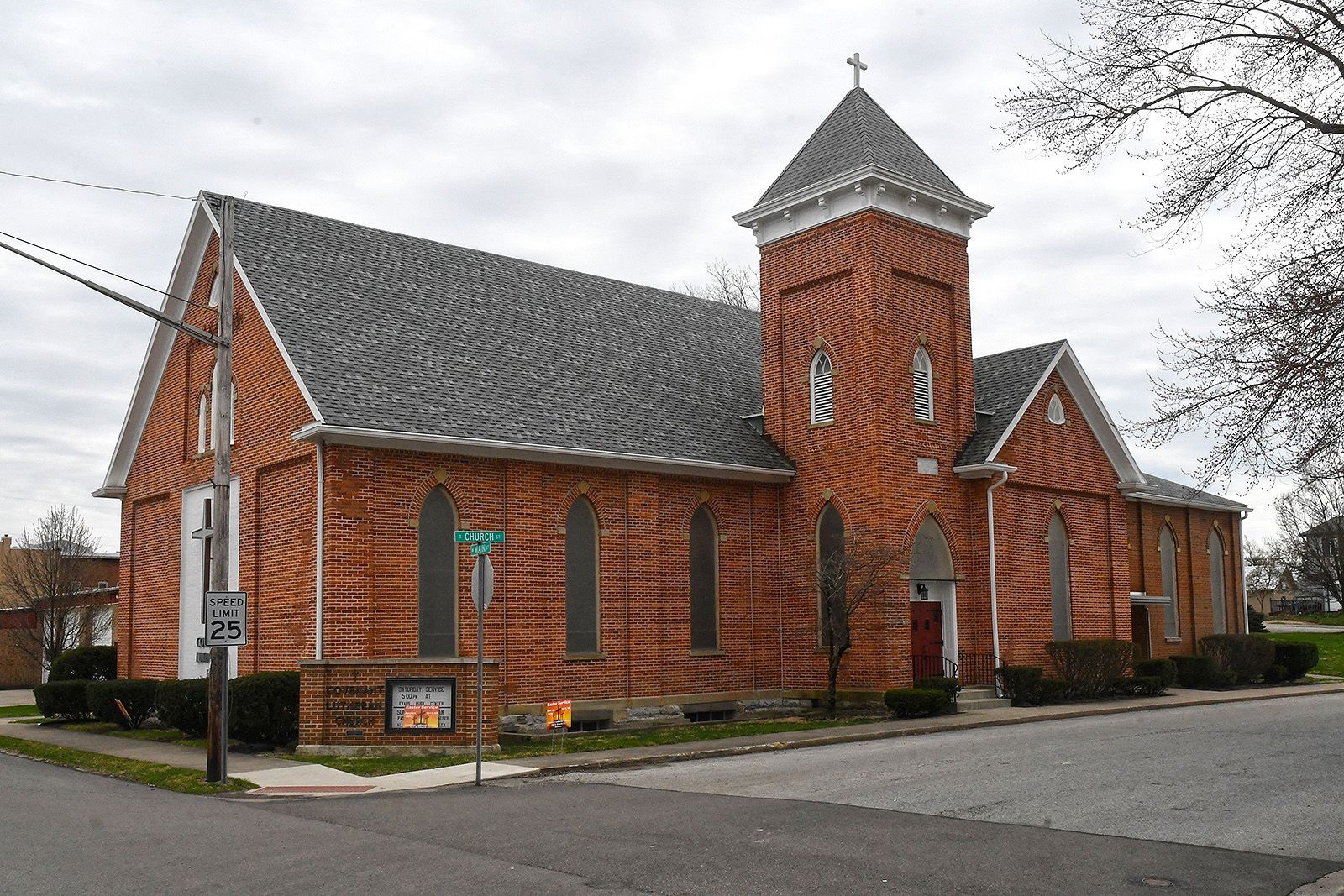 Brick church with a steeple and cross. Located on a street corner, gray roof.