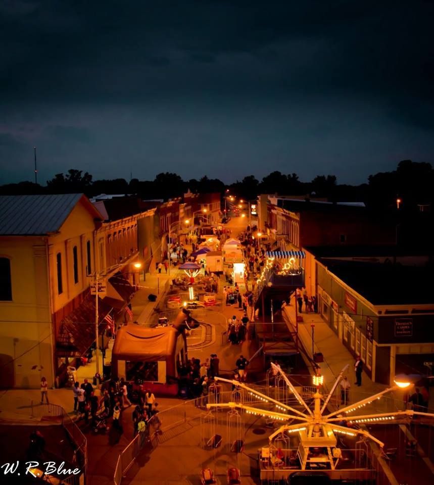 Nighttime street fair with illuminated buildings, crowds, and a small carnival ride.