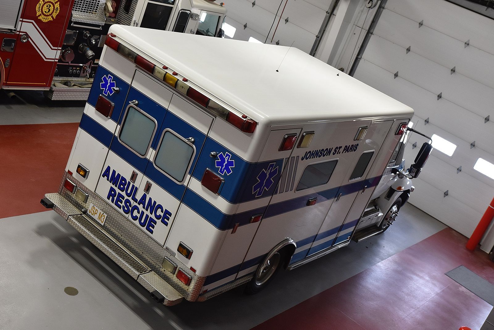 Ambulance parked in a garage. White with blue stripes and emergency lights flashing, chrome accents. 