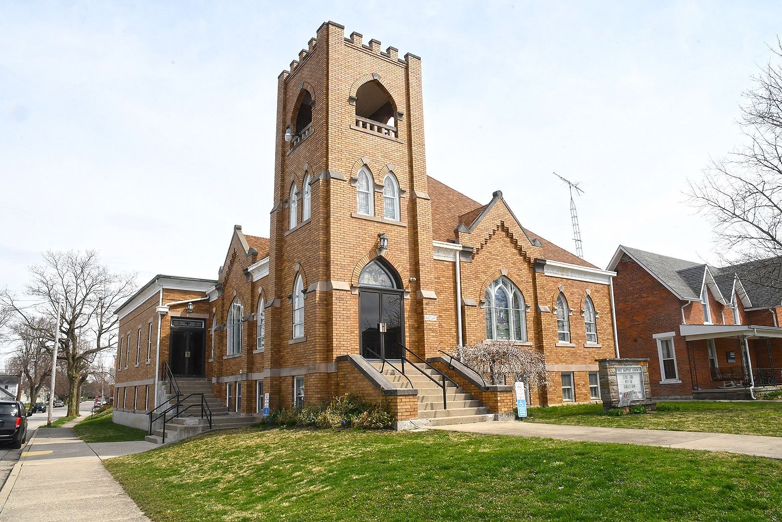 Brick church building with a tall tower and arched windows, set on a grassy lawn with a sidewalk.