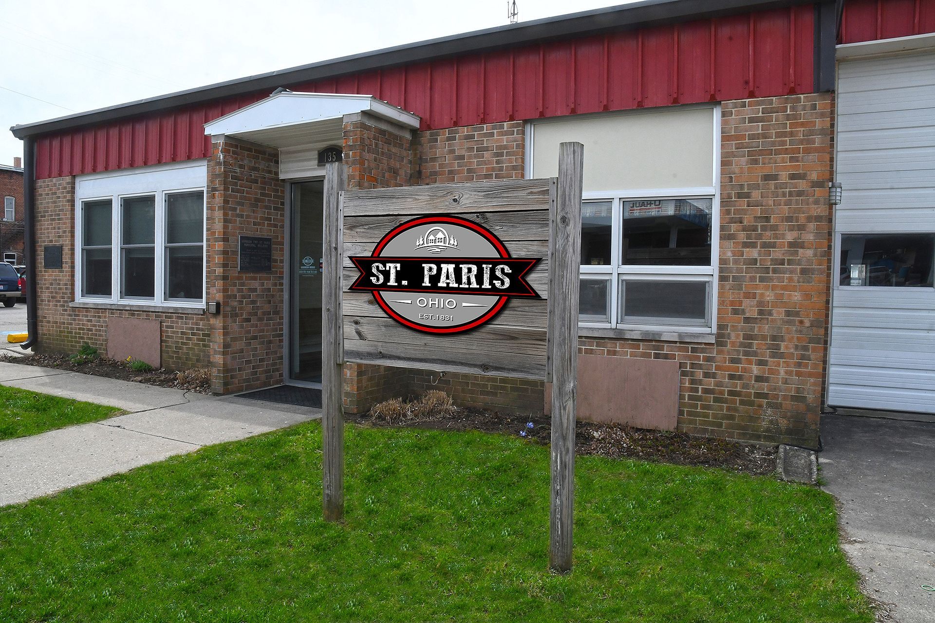 Sign for St. Paris in front of a brick building with a red roof. Green lawn in front.