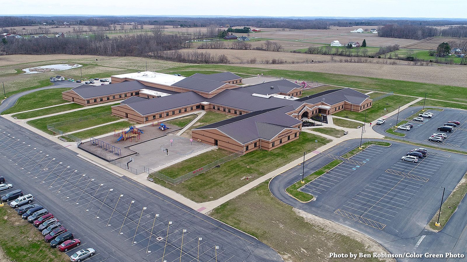 Aerial view of a one-story brick school building with a playground, parking lots, and surrounding fields.