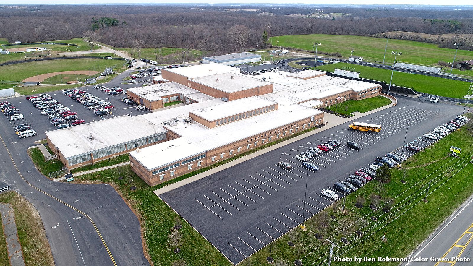 Aerial view of a large, brown brick school building with multiple parking lots and a yellow school bus.