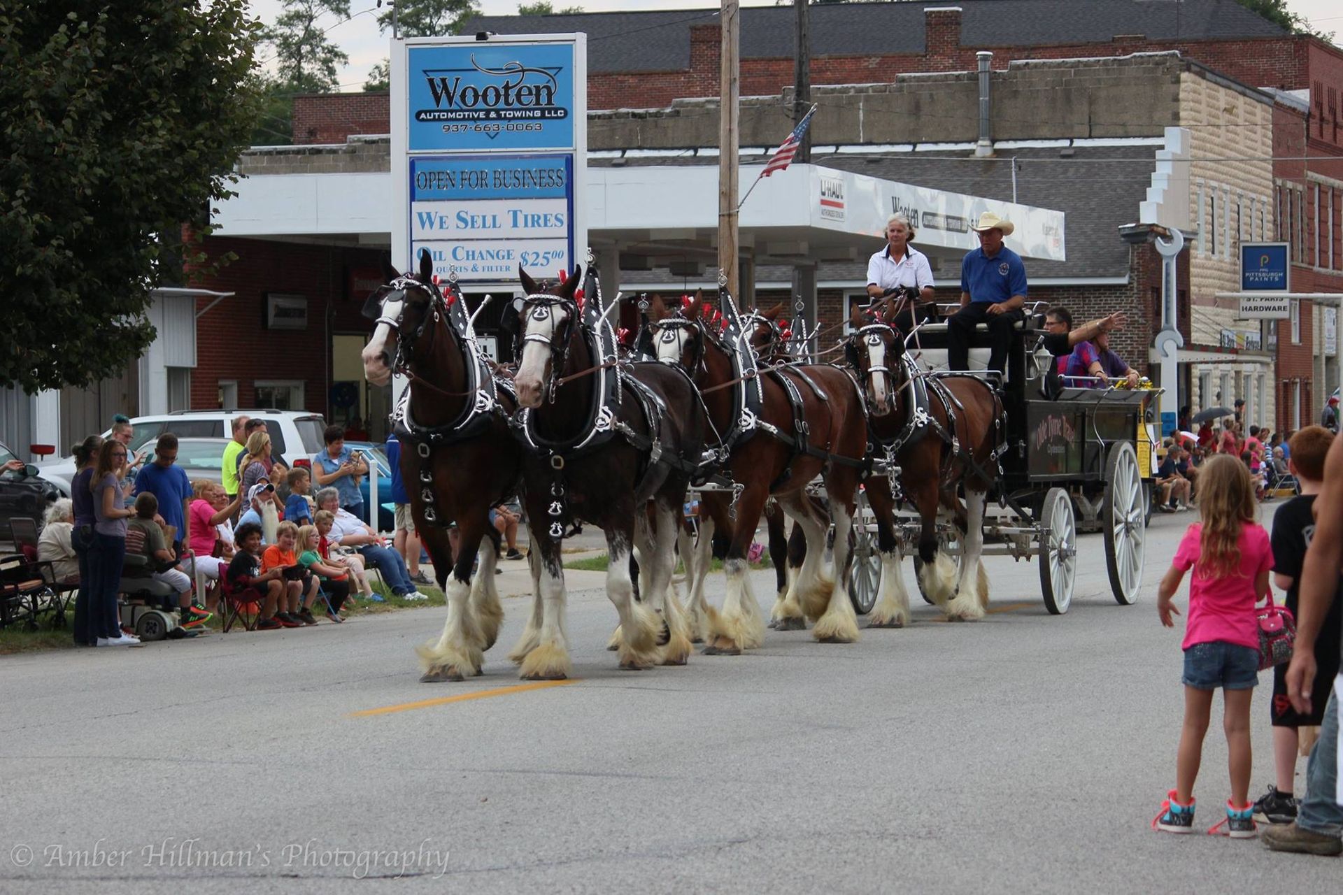 Eight Clydesdale horses pulling a wagon in a parade; onlookers watch.