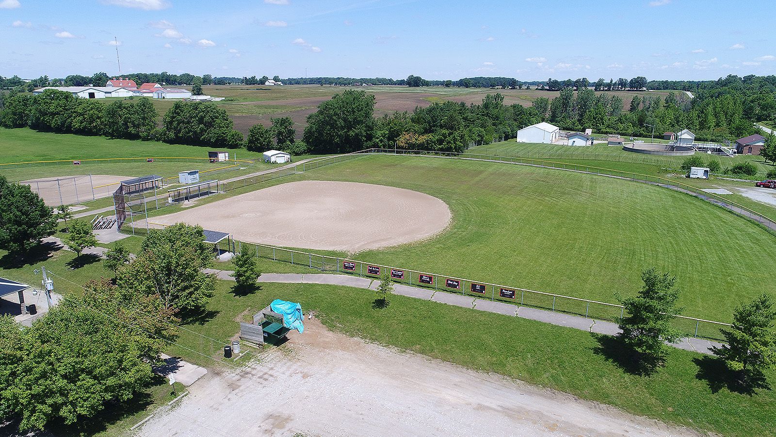 Aerial view of a green field with a dirt area, trees, and buildings in the background on a sunny day.