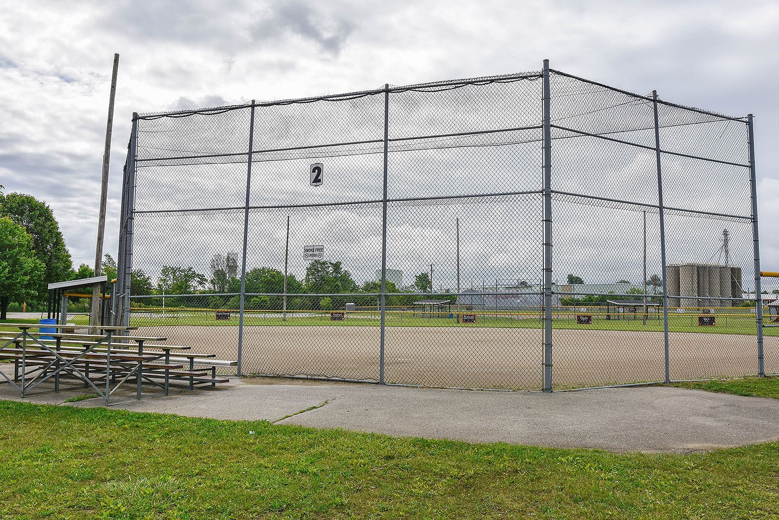 A baseball batting cage outdoors, with netting and a gravel base, stands on grass under an overcast sky.