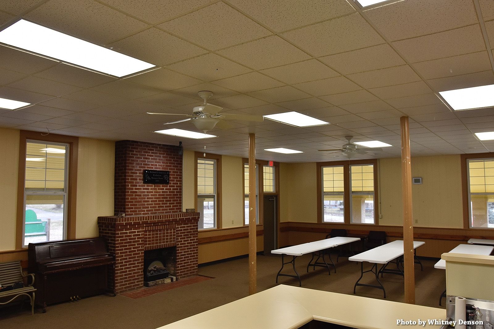 Interior of a community center with tables, fireplace, and windows, lit by fluorescent lights.