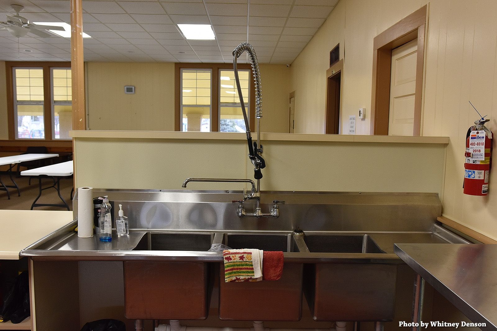 Stainless steel triple sink in a kitchen, with a tall faucet and a fire extinguisher on the wall.
