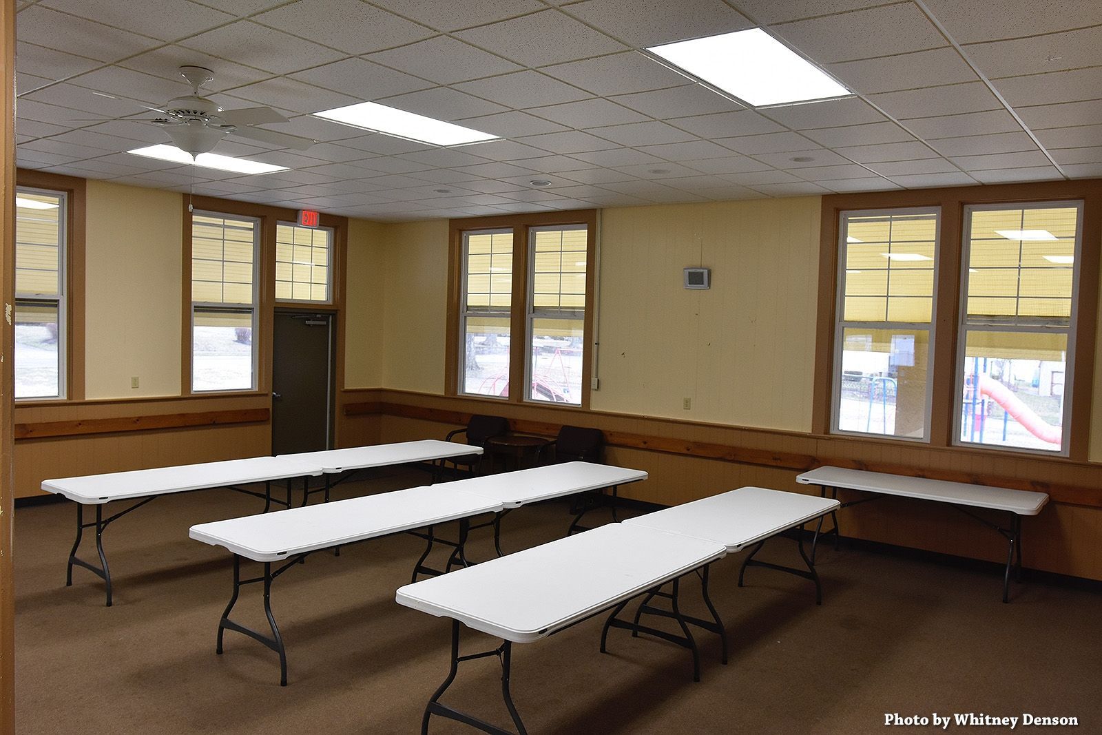 Empty room with rectangular tables, windows, beige walls, and brown trim.