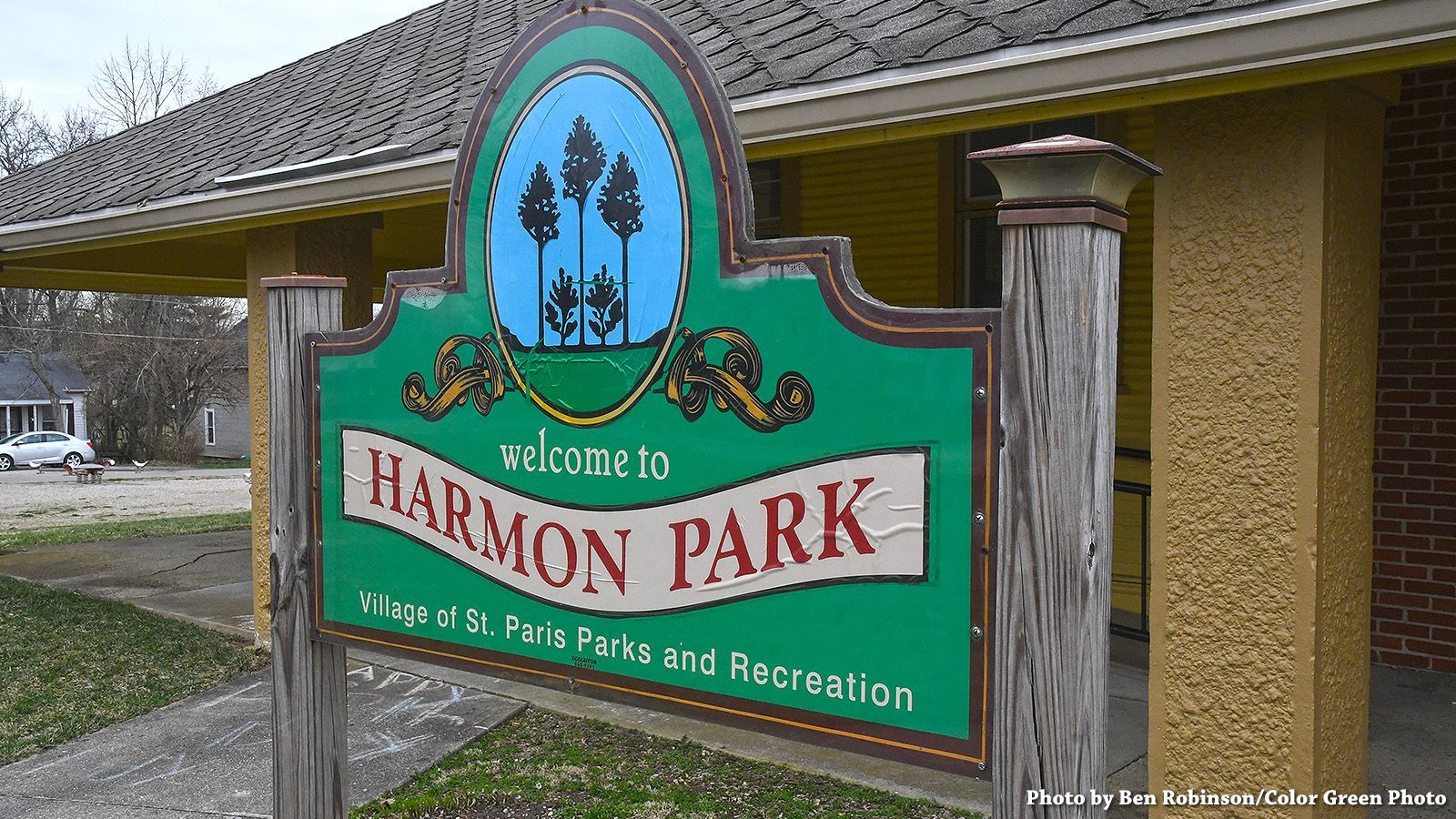 Sign for Harmon Park, green with white text, illustration of trees, wooden posts, building background.
