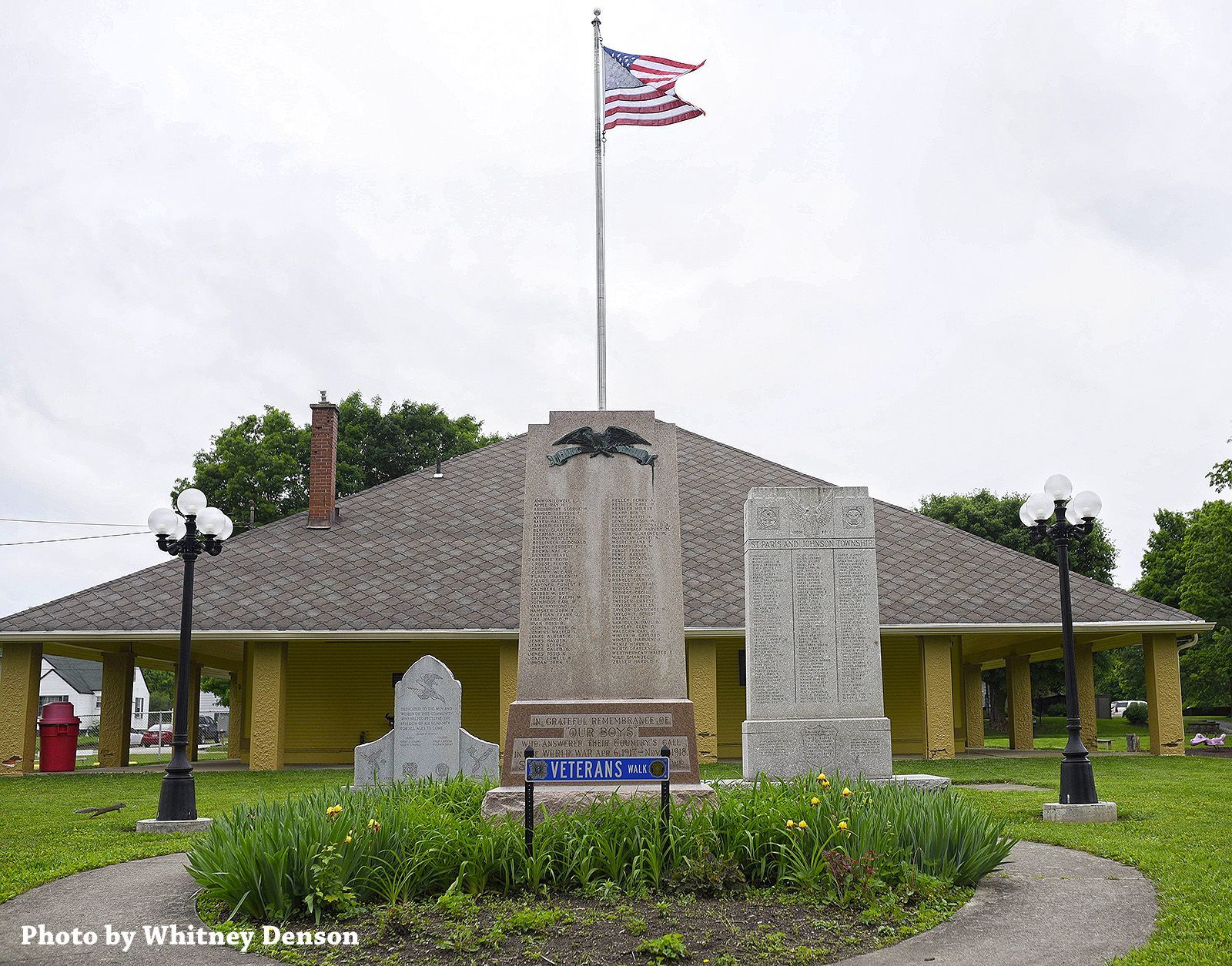 War memorial with flag, names, and a yellow building with a dark roof.