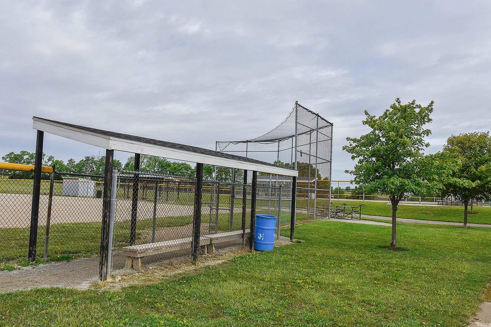 A baseball batting cage and backstop on a grassy field under a cloudy sky.