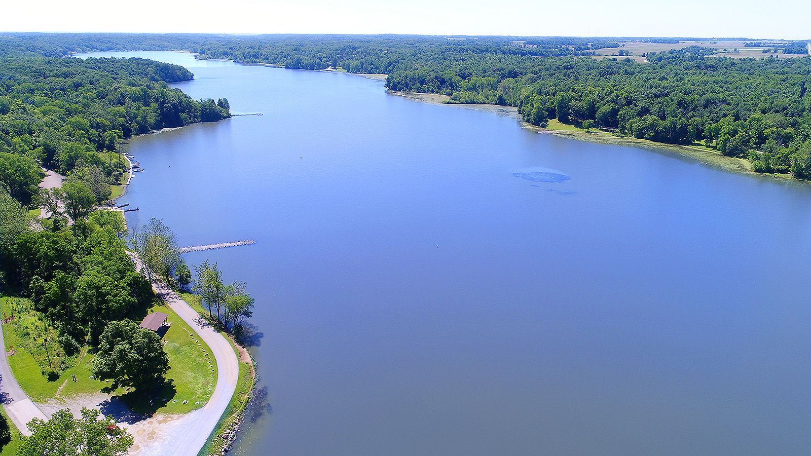 Wide aerial view of a lake bordered by green trees and a park path, with a blue sky overhead.