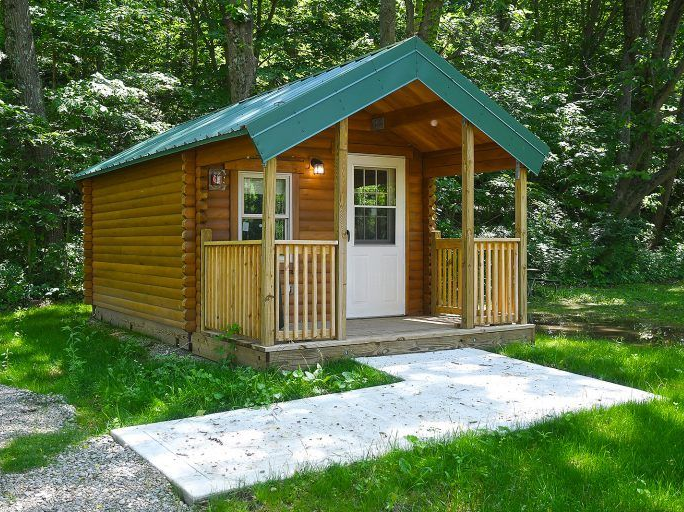 Small wooden cabin with green roof and porch, set in a grassy area with trees.