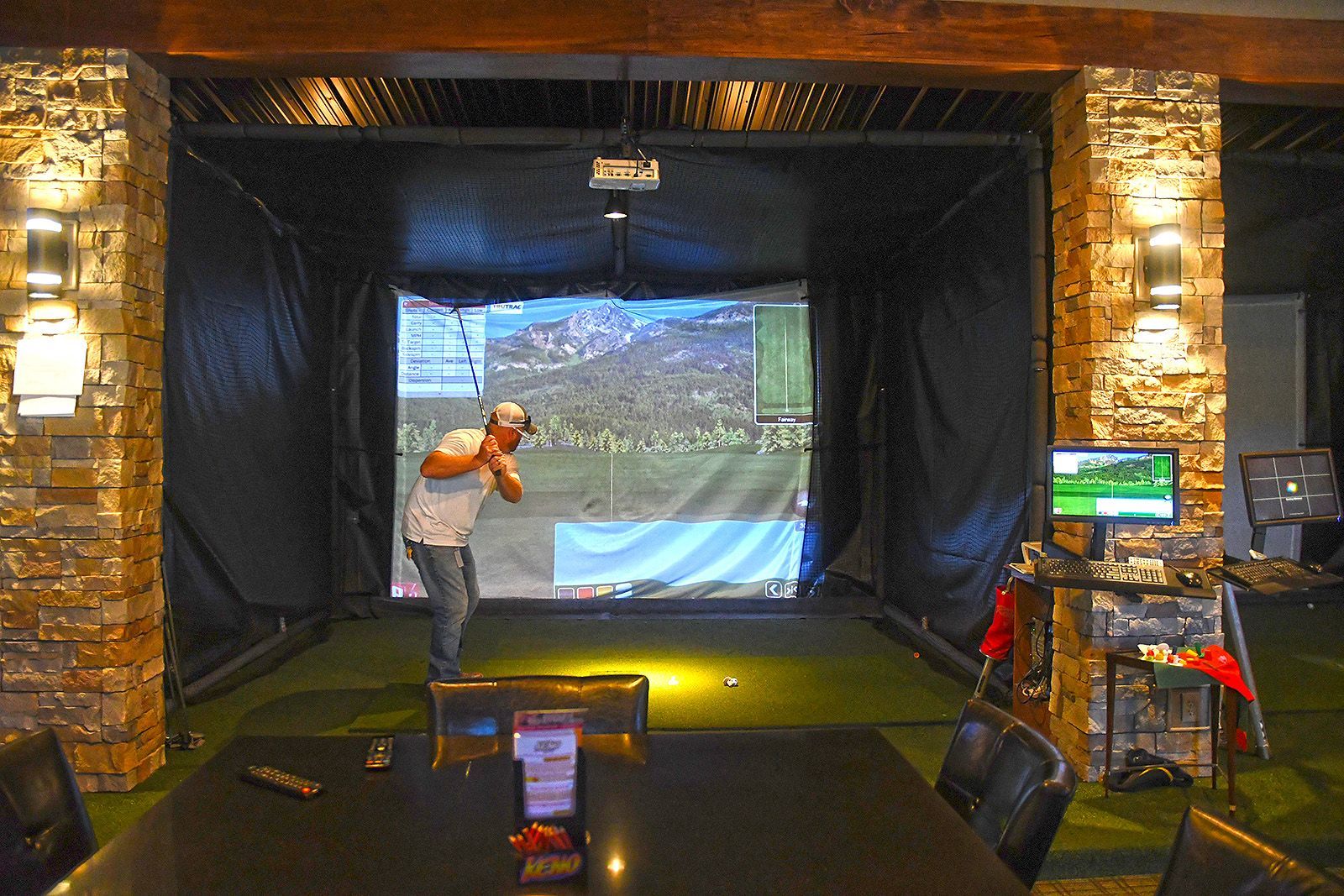 Man golfing in a golf simulator. Dark enclosure with screen displaying course. Brown stone columns, golf ball on green turf.