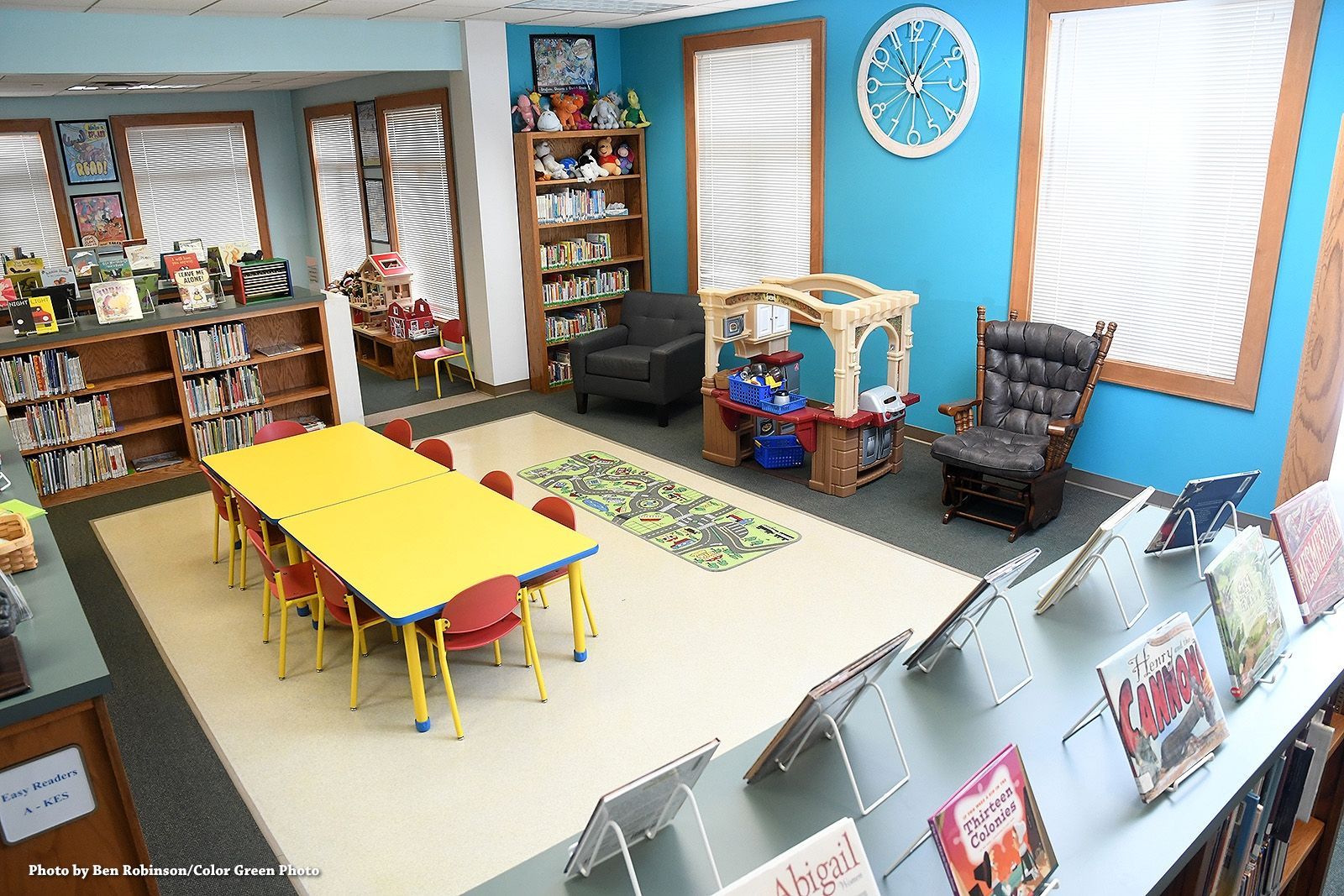 Children's library room: yellow table with chairs, bookshelves, playhouse, blue walls, a clock, and windows.